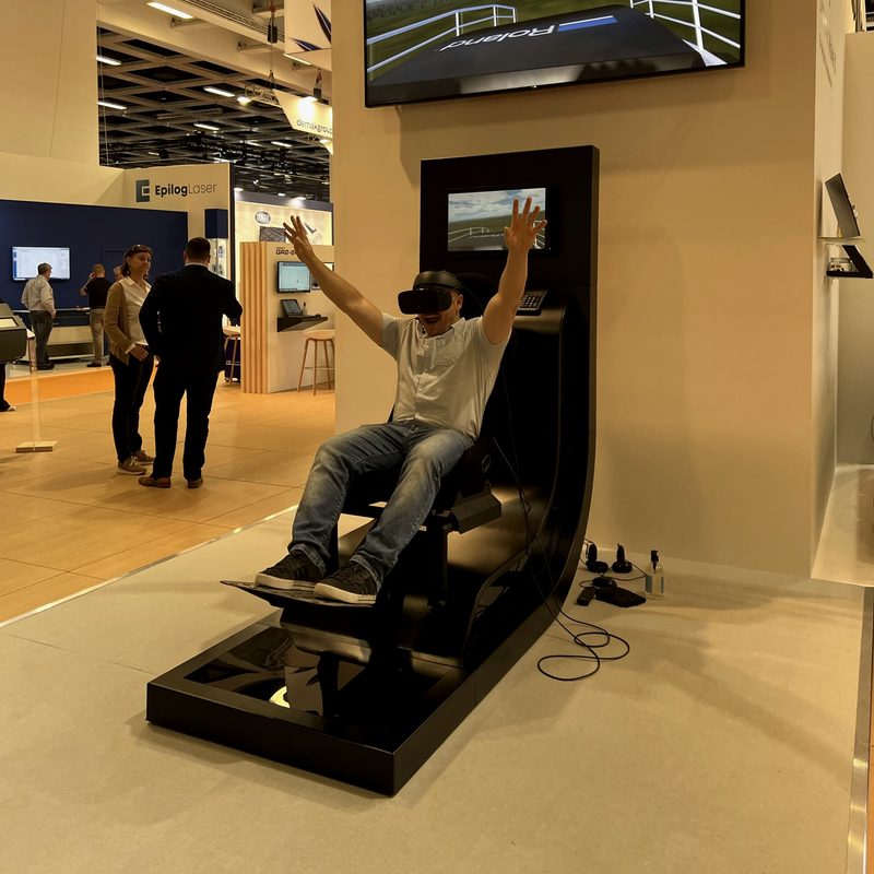 A rider in a VR headset with arms raised on the full-motion roller coaster, the ride shown on a large screen above