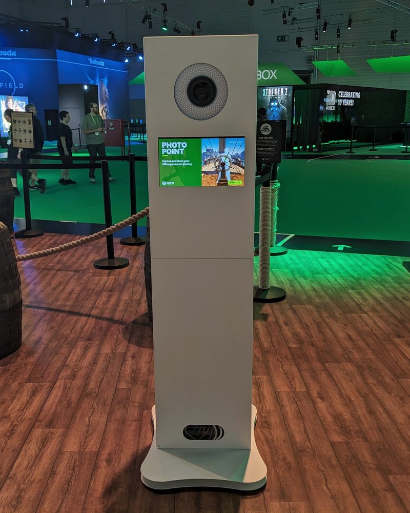 A free-standing white photobooth tower with a ring-lit camera and touchscreen on an exhibition stand